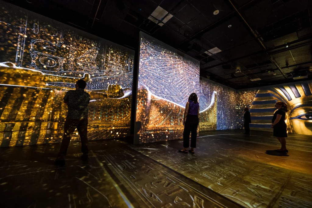 Multiple people standing in a darkened room with Egyptian imagery projected on one wall and floor and a portion of King Tutankhamen's gold death mask on another wall.